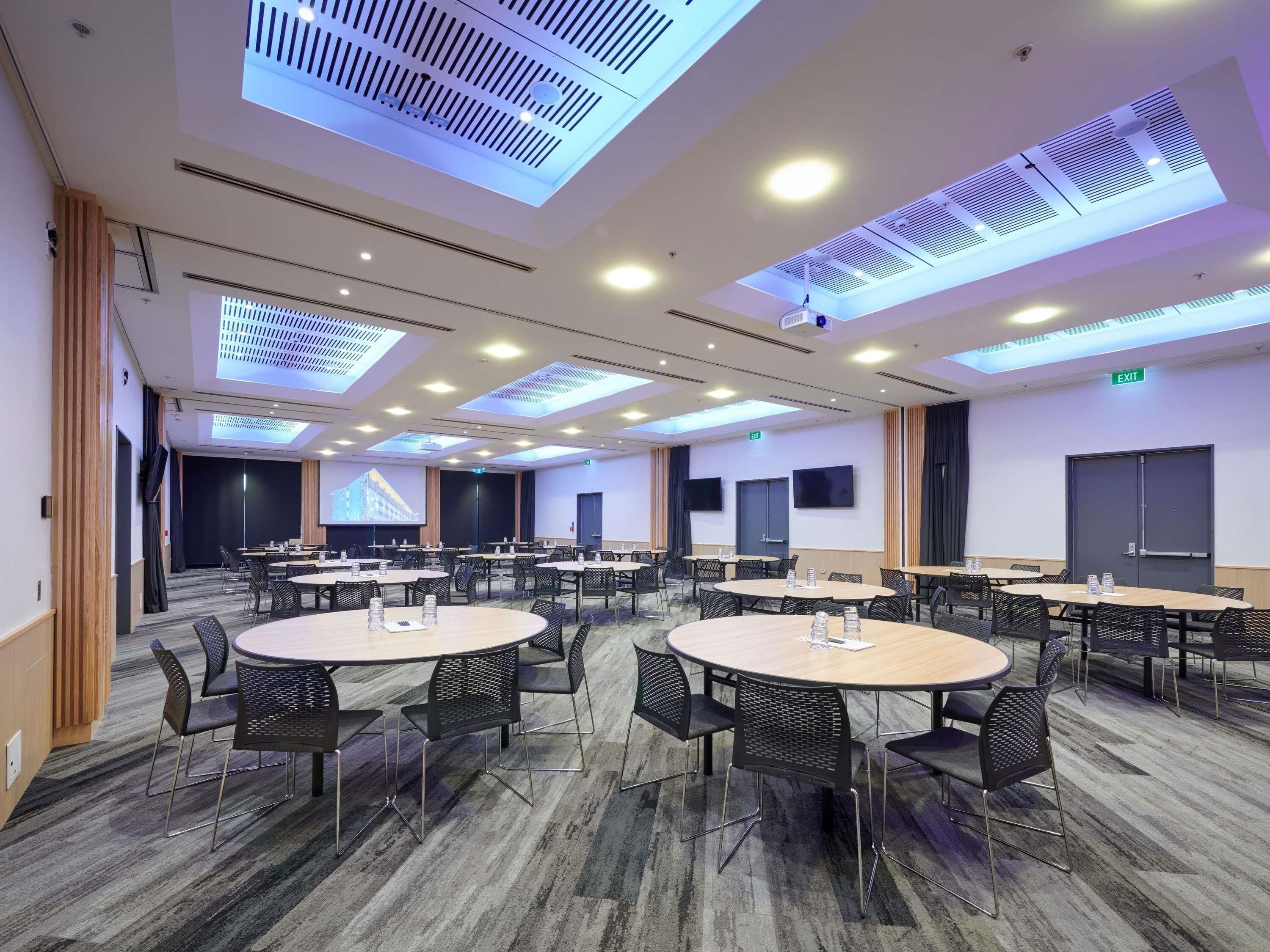 Un salón de conferencias con mesas y sillas redondas, una pantalla grande y un techo con tragaluces.