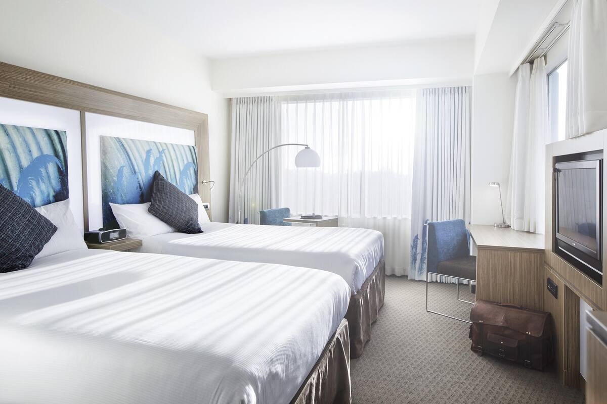 A hotel room with two beds, a desk, a chair, a TV, and a suitcase.