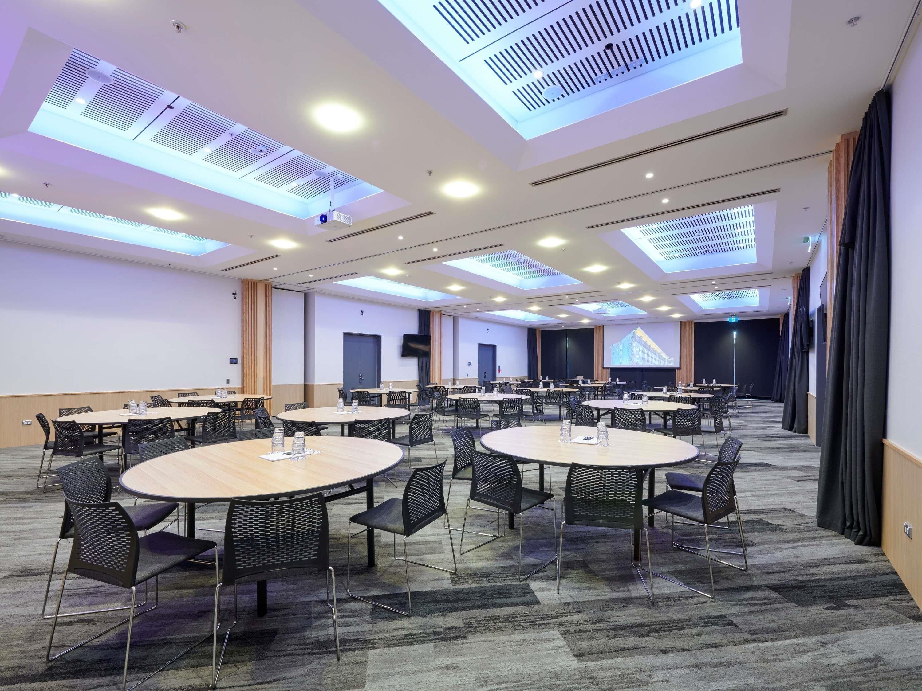 A conference room with multiple tables and chairs, a projector screen, and a large screen displaying a building image.
