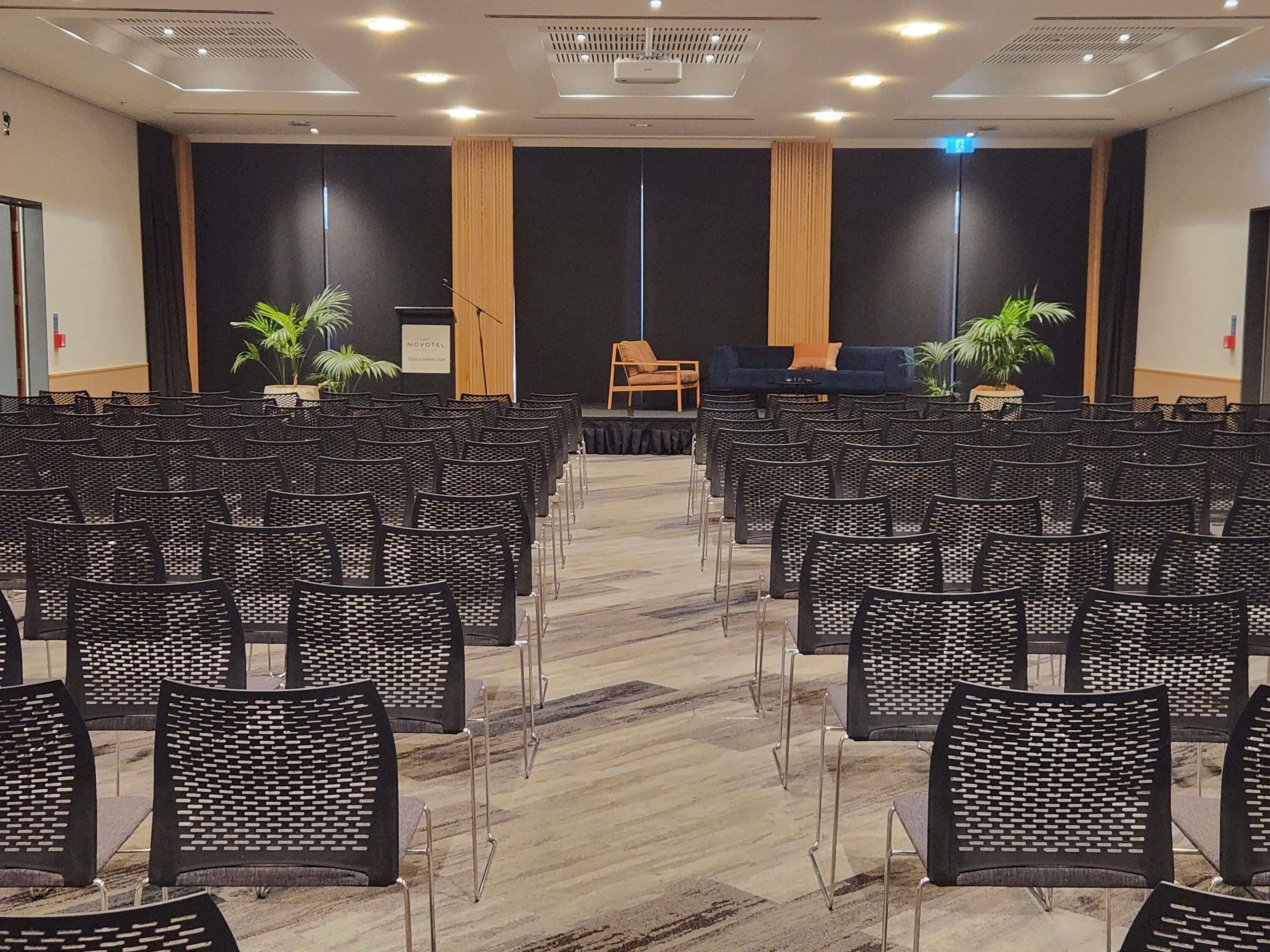 A conference room with rows of chairs, a stage with a podium, and two potted plants.