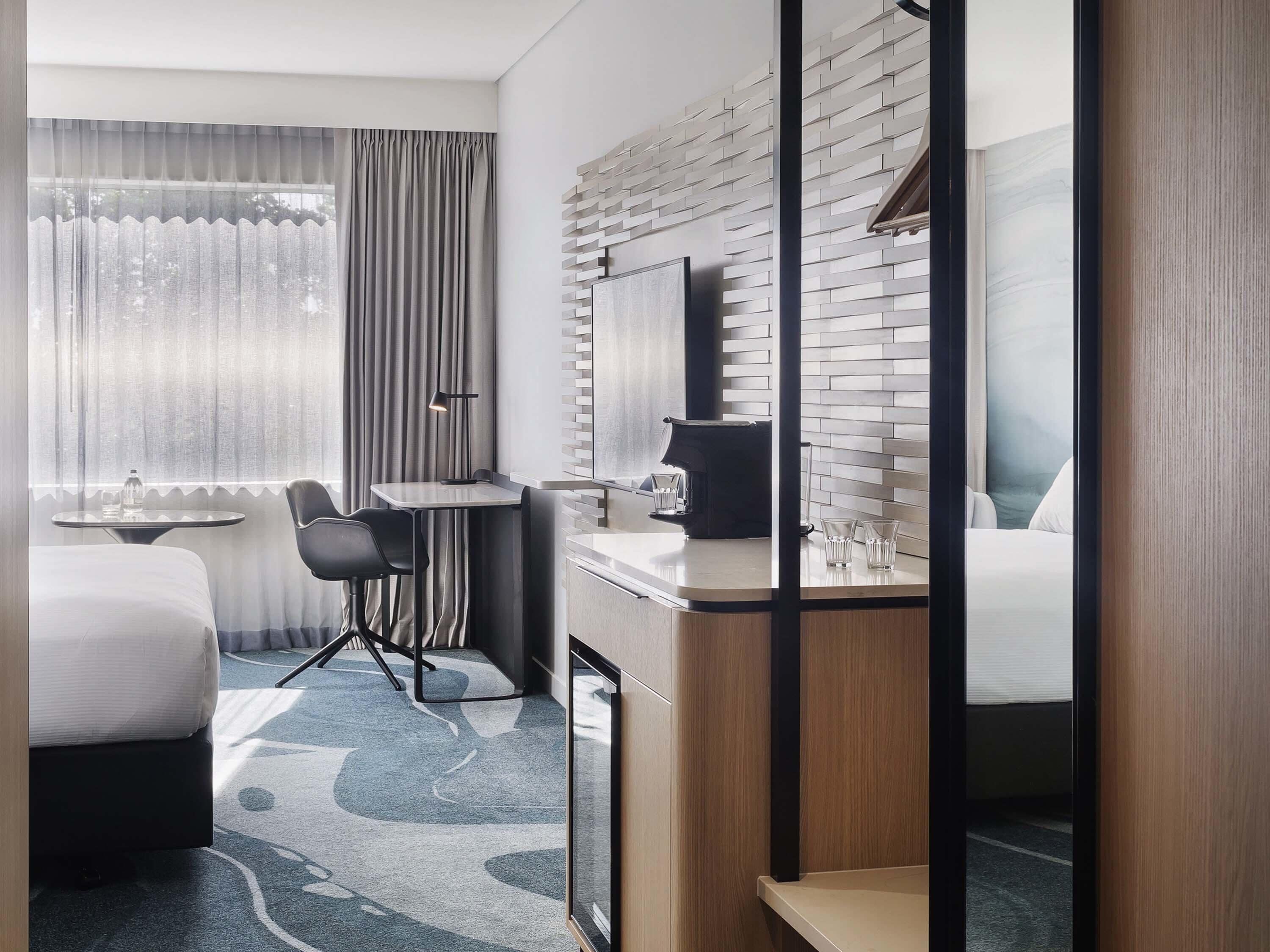 A hotel room with a bed, a desk with a chair, a small table, a mirror, and a window with curtains.
