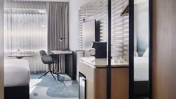 A hotel room with a bed, a desk with a chair, a small table, a mirror, and a window with curtains.