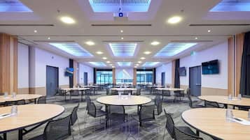 Un salón de conferencias con varias mesas y sillas, luces en el techo y una pantalla grande.