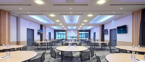 Un salón de conferencias con varias mesas y sillas, luces en el techo y una pantalla grande.