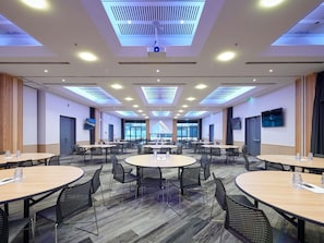 Un salón de conferencias con varias mesas y sillas, luces en el techo y una pantalla grande.
