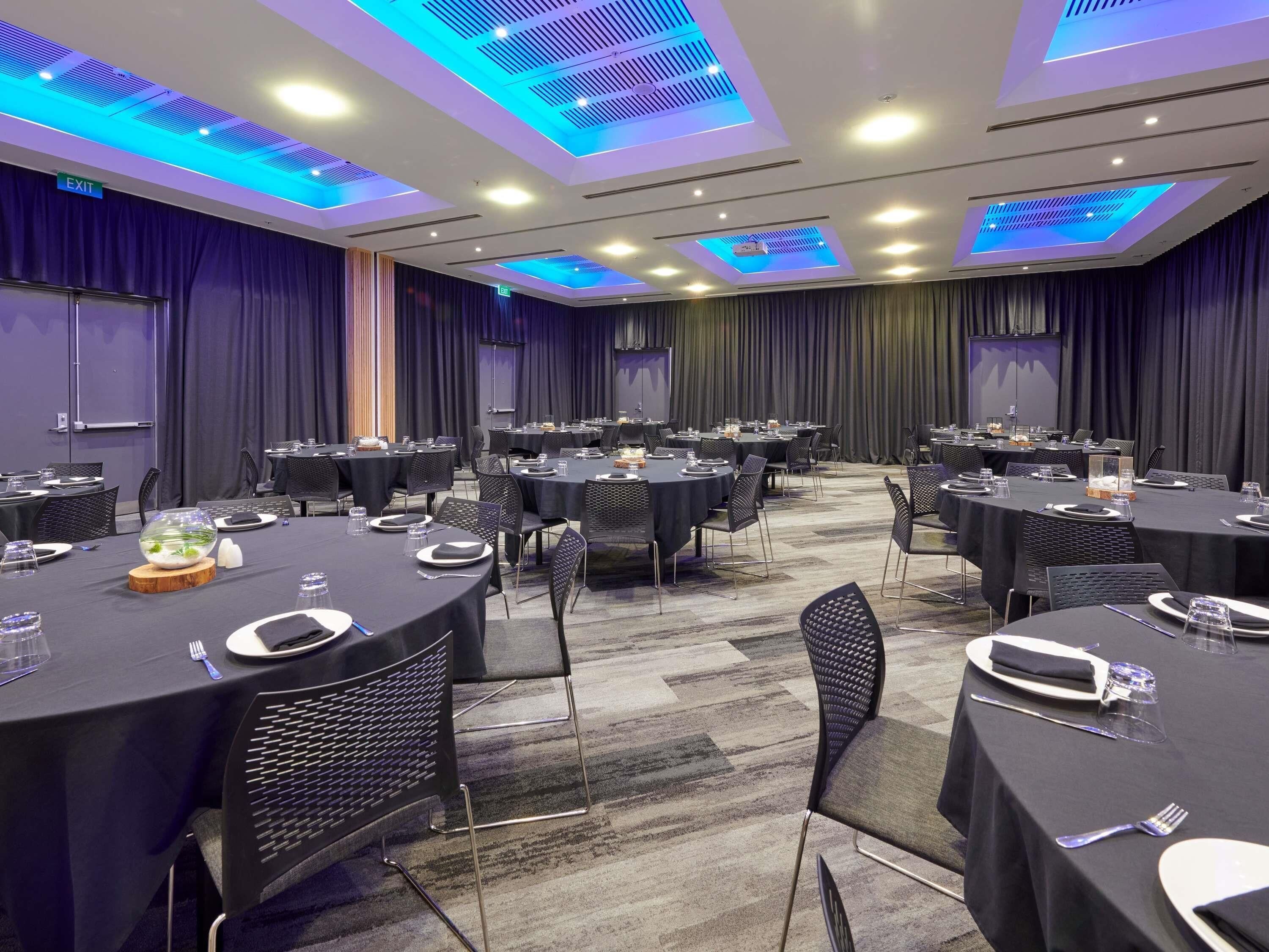 Un salón de conferencias con mesas redondas, sillas negras e iluminación azul en el techo.