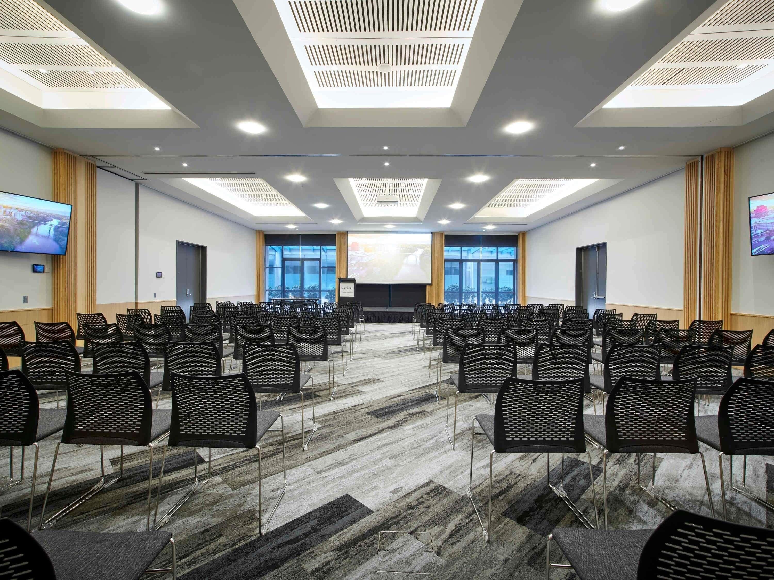 Un salón de conferencias con filas de sillas, una pantalla grande y un proyector.