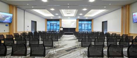 Un salón de conferencias con filas de sillas, una pantalla grande y un proyector.