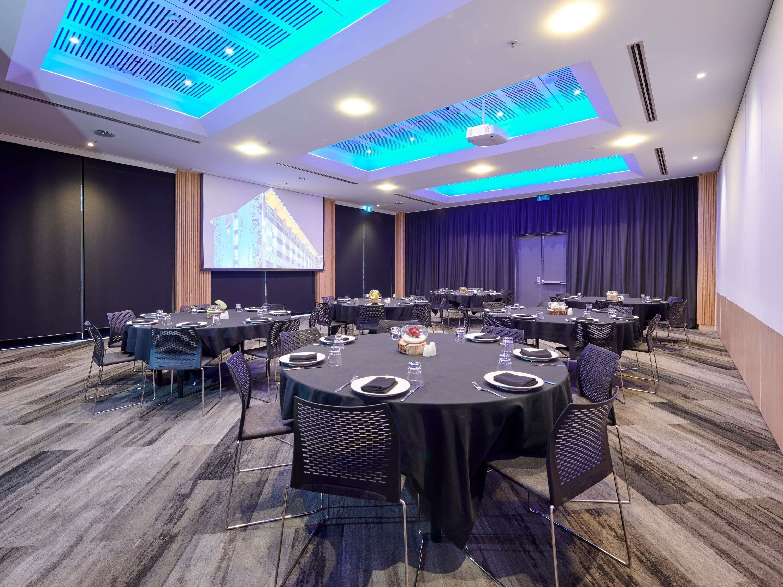 Un salón de conferencias con mesas redondas, sillas, una pantalla de proyector y una pantalla para presentaciones.