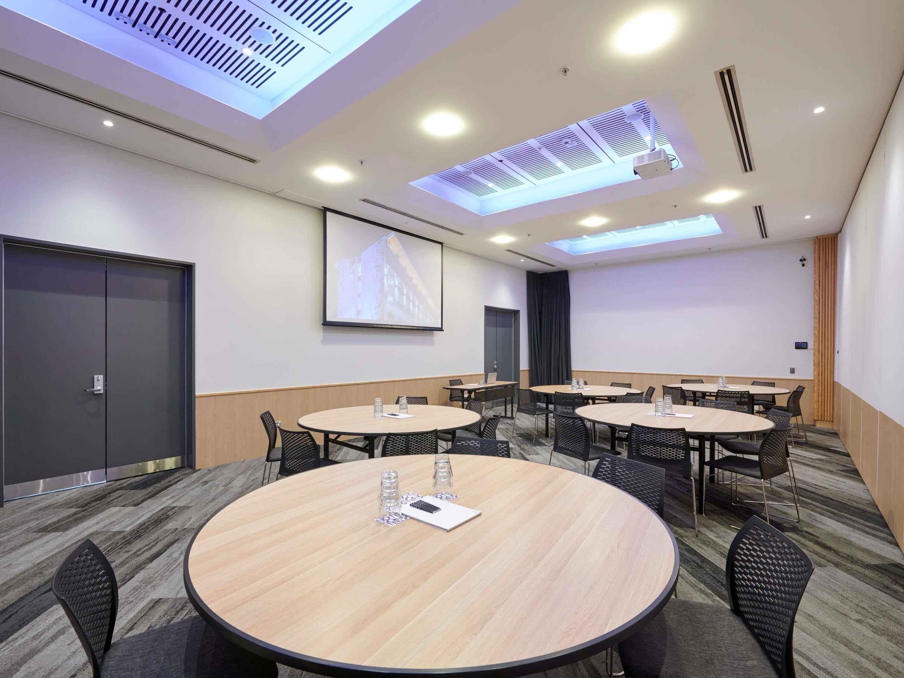 A conference room with multiple tables, chairs, a projector screen, and a large window with skylights.