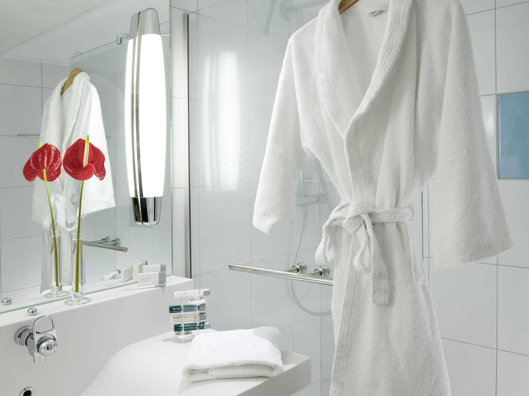 A modern bathroom with a white bathtub, a vanity with toiletries, a glass shower enclosure, and a white bathrobe hanging on a rack.