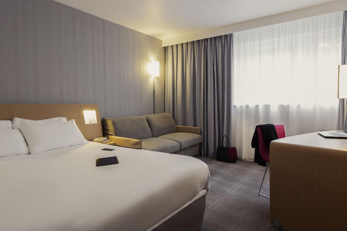 A hotel room with a large bed, a sofa, a desk with a laptop, and a window with curtains.