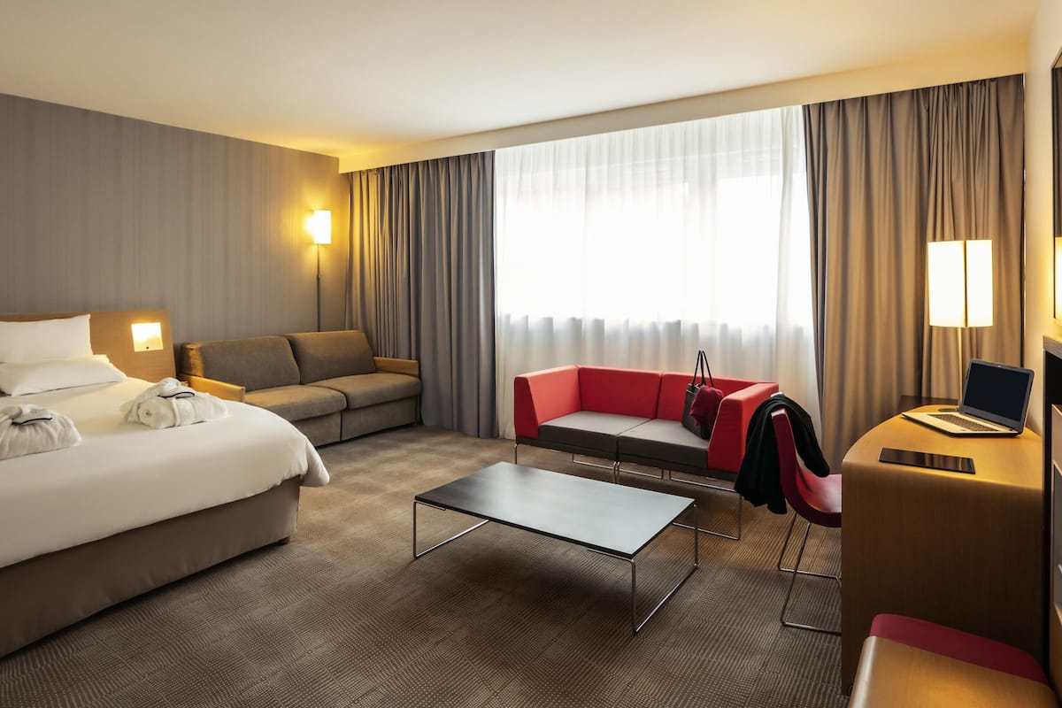 A hotel room with a bed, two sofas, a desk with a laptop, a lamp, and a large window with curtains.