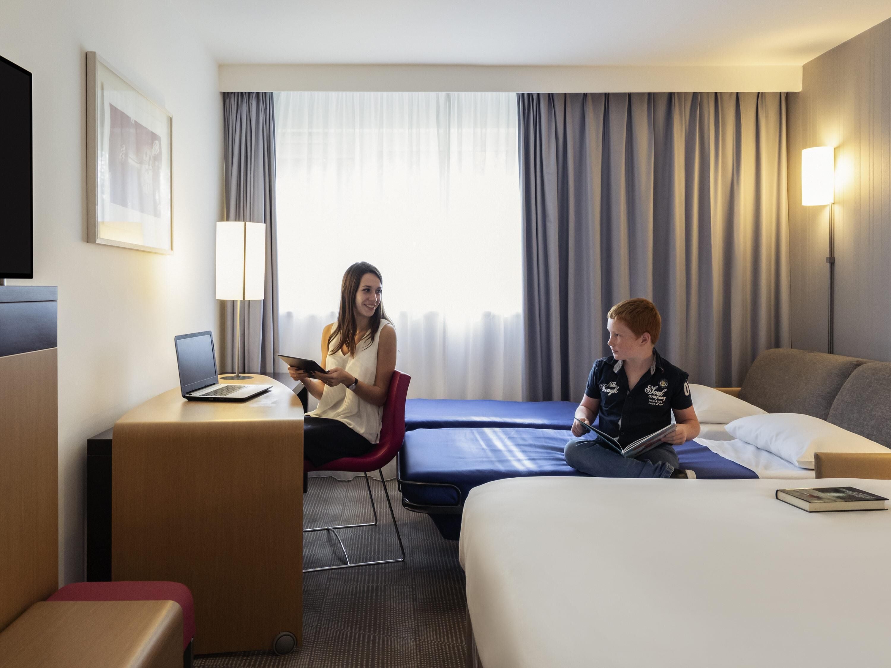 A hotel room with a desk, two beds, a TV, and a window with curtains.