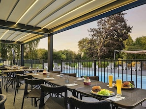 An outdoor dining area with tables and chairs, a pool, and a view of trees and a car in the background.