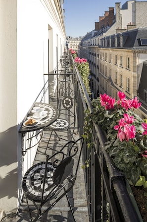 Un balcón con mesa y sillas, una planta en maceta con flores, y vistas a edificios.