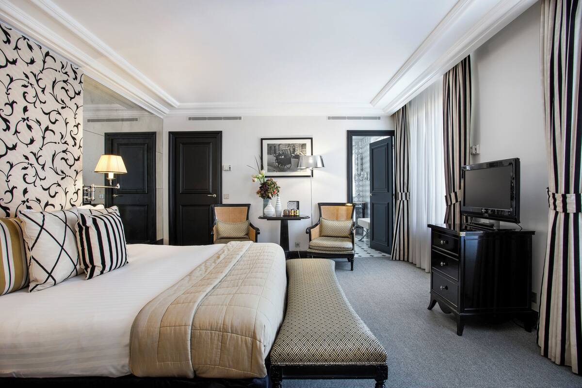 Habitación de hotel con una cama grande, dos sillones, una mesita con una lámpara, televisión y un cuadro en la pared.