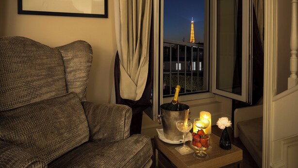 A room with a view of the Eiffel Tower at night, a cozy seating area with a patterned sofa, a small table with a champagne bottle, glasses, and candles.