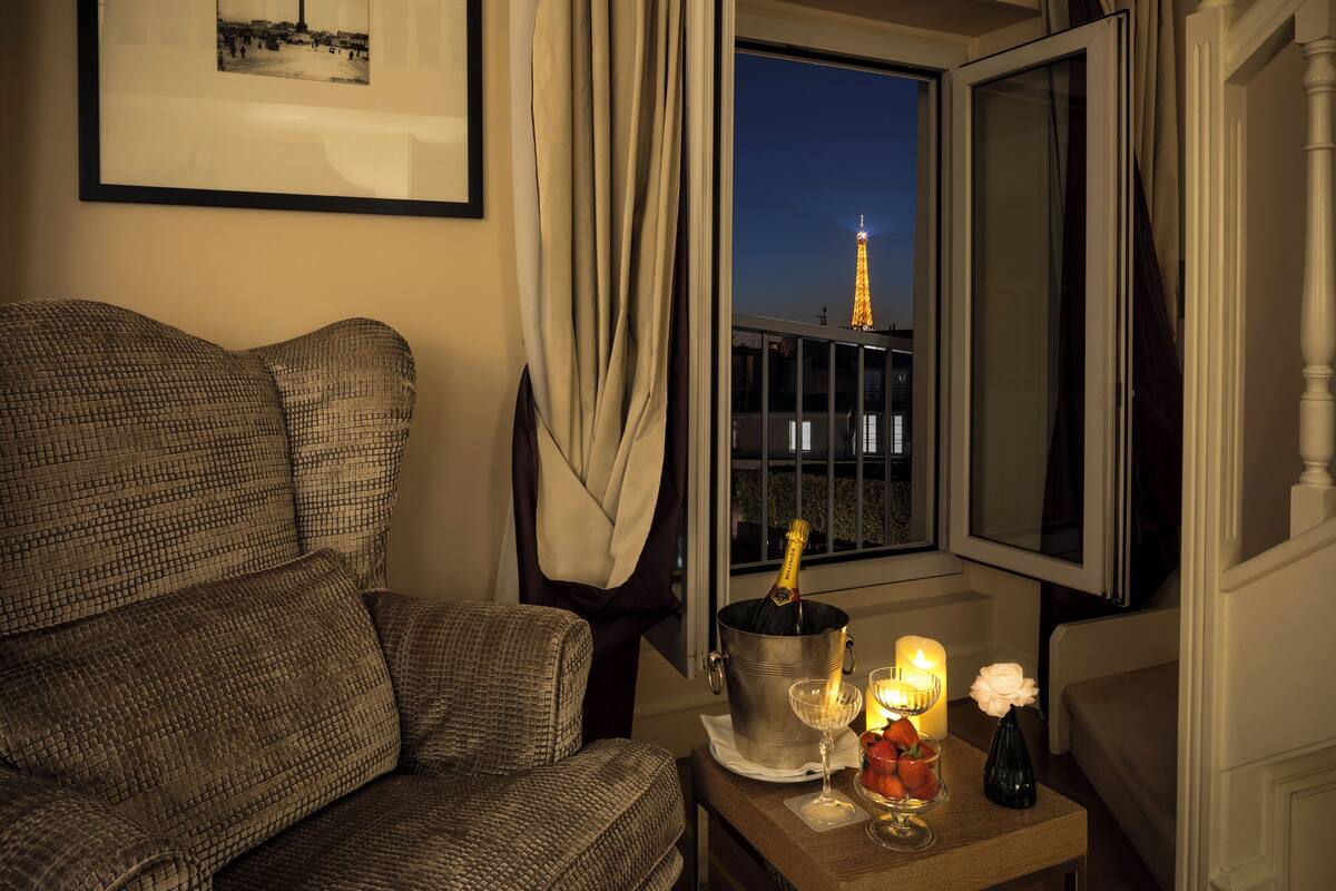 Una habitación con vistas a la Torre Eiffel de noche, una zona de estar acogedora con un sofá estampado, una mesita con una botella de champán, copas y velas.