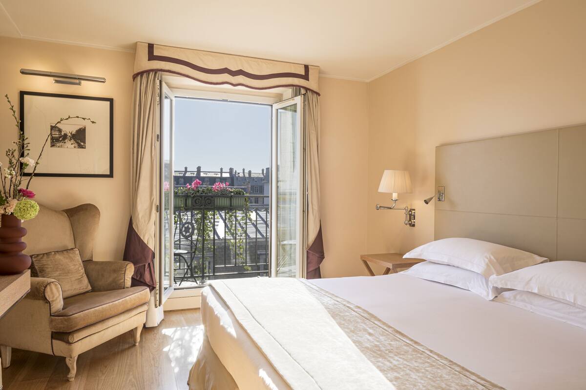 Una habitación de hotel con cama, una silla, mesita de noche, lámpara y ventana con cortinas.