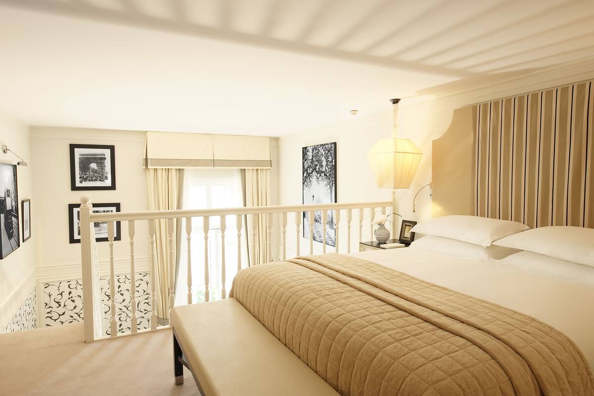 Habitación de hotel con una cama grande, un banco, una escalera, vistas al exterior y una lámpara.