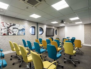 Une salle de conférence au design moderne, équipée d’un grand téléviseur à écran plat, d’un projecteur et d’un ensemble de chaises aux couleurs vives.