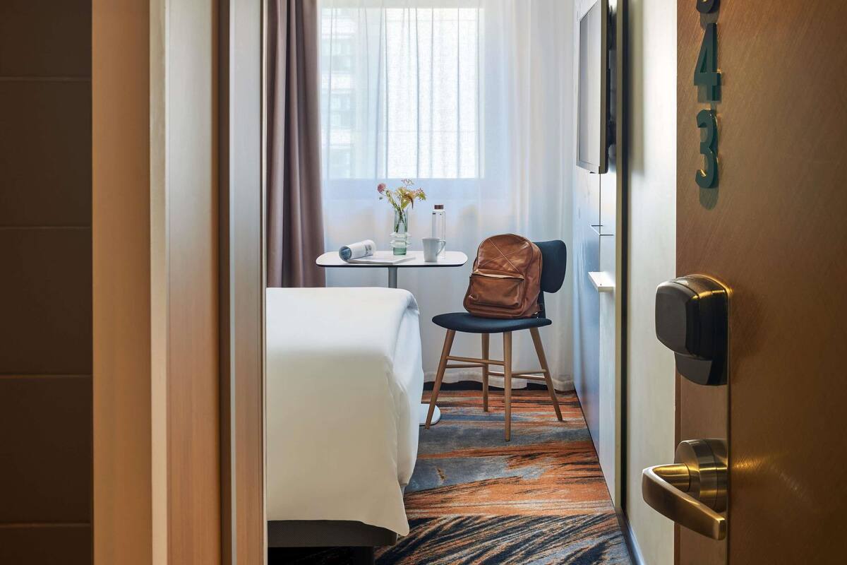 Une chambre d’hôtel avec un lit, une chaise, une petite table sur laquelle se trouve un vase, et une porte avec un numéro.