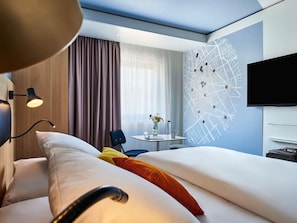 A hotel room with a bed, a desk, a chair, a TV, and a wall with a map design.