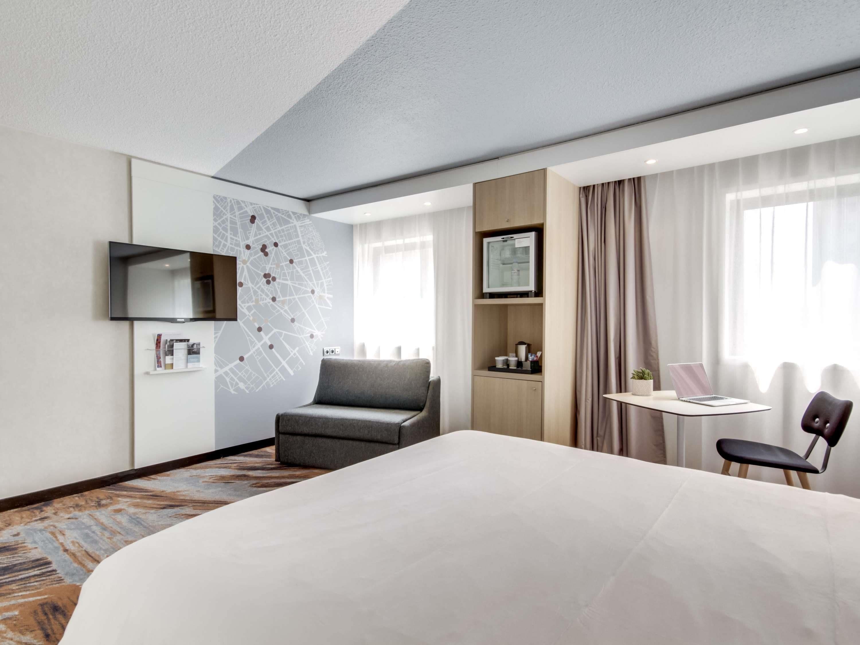 Una camera d'albergo con un letto, una scrivania, una sedia, un televisore, una mappa appesa al muro e una finestra con tende.