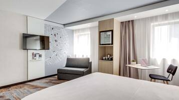 Una camera d'albergo con un letto, una scrivania, una sedia, un televisore, una mappa appesa al muro e una finestra con tende.