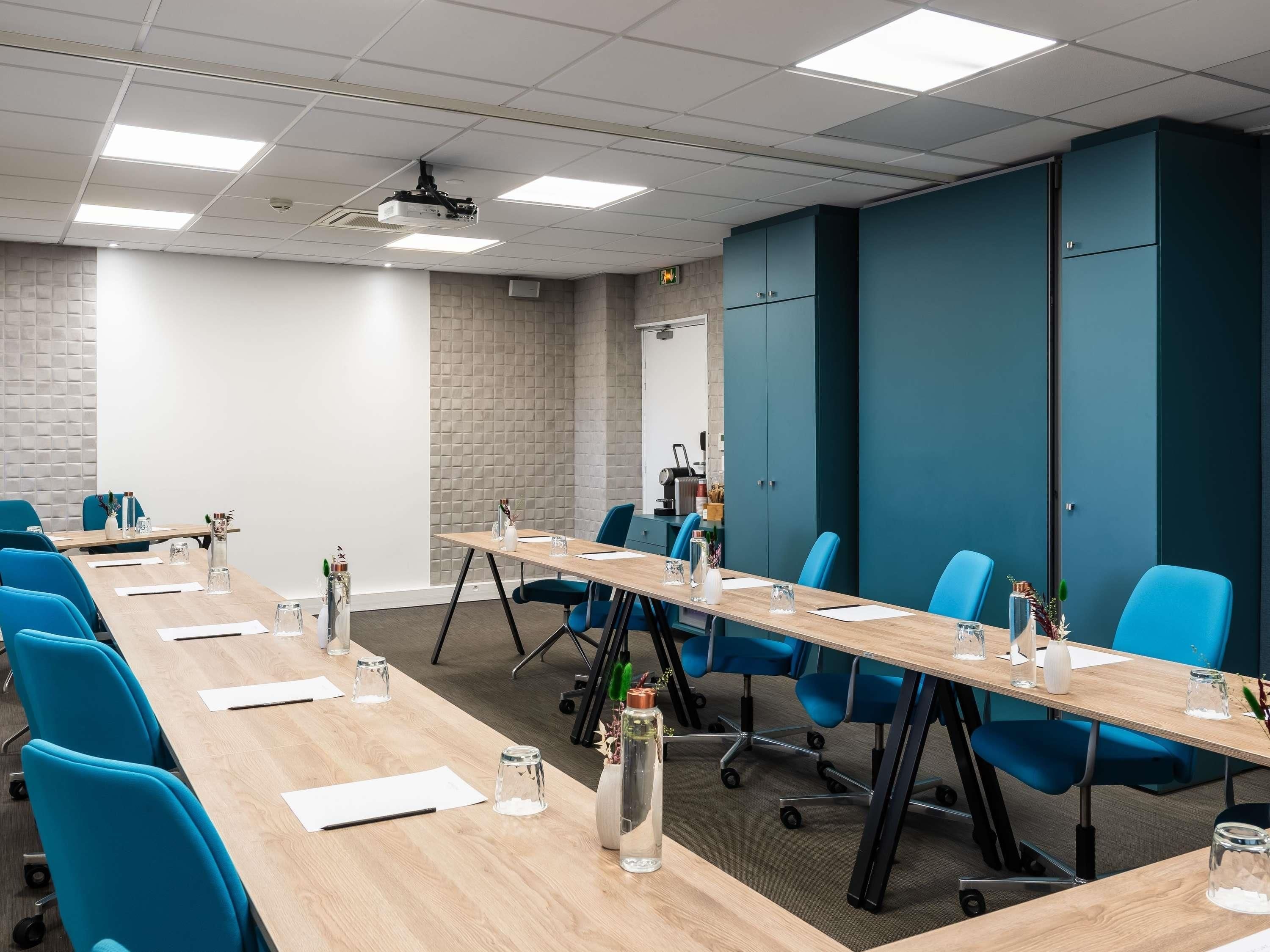 Un salón de conferencias con una larga mesa, sillas azules y un proyector en el techo.