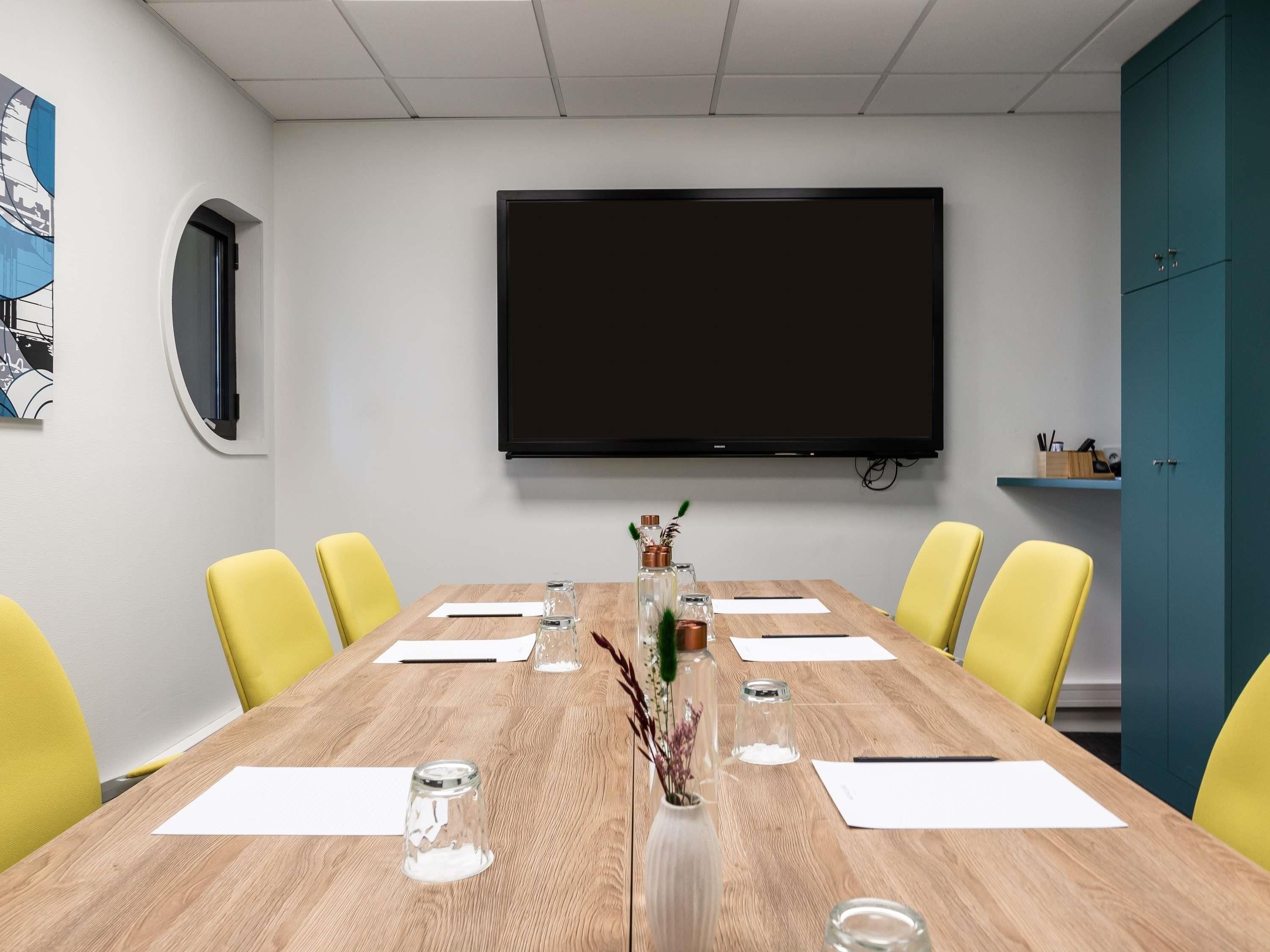 A conference room with a large flat-screen TV, wooden table, yellow chairs, and a teal cabinet.