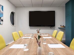 Een vergaderruimte met een groot flatscreen televisietoestel, een houten tafel, gele stoelen en een blauwgroene kast.