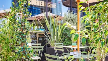An outdoor seating area with tables and chairs, surrounded by greenery and a building with a 'CORNER TATION' sign.