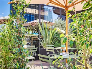 Un área de estar al aire libre con mesas y sillas, rodeada de follaje y un edificio con un letrero que dice 'CORNER TATION'.