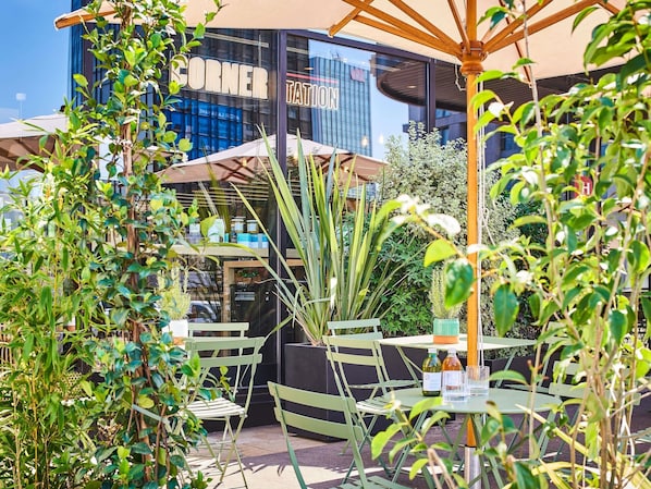 An outdoor seating area with tables and chairs, surrounded by greenery and a building with a 'CORNER TATION' sign.