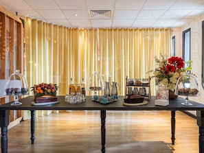 Une chambre d’hôtel avec une longue table dressée, sur laquelle on trouve divers objets, notamment une présentation de fruits, des bouteilles et un arrangement floral.