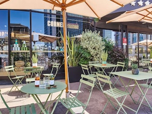 An outdoor seating area with tables and chairs, a large umbrella, and a view of a building with the sign 'CORNER STATION'.