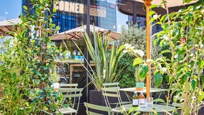 Een terras met groene stoelen en parasols, een glazen gevel met 'CORNER TATION' aanduiding, en verschillende planten.