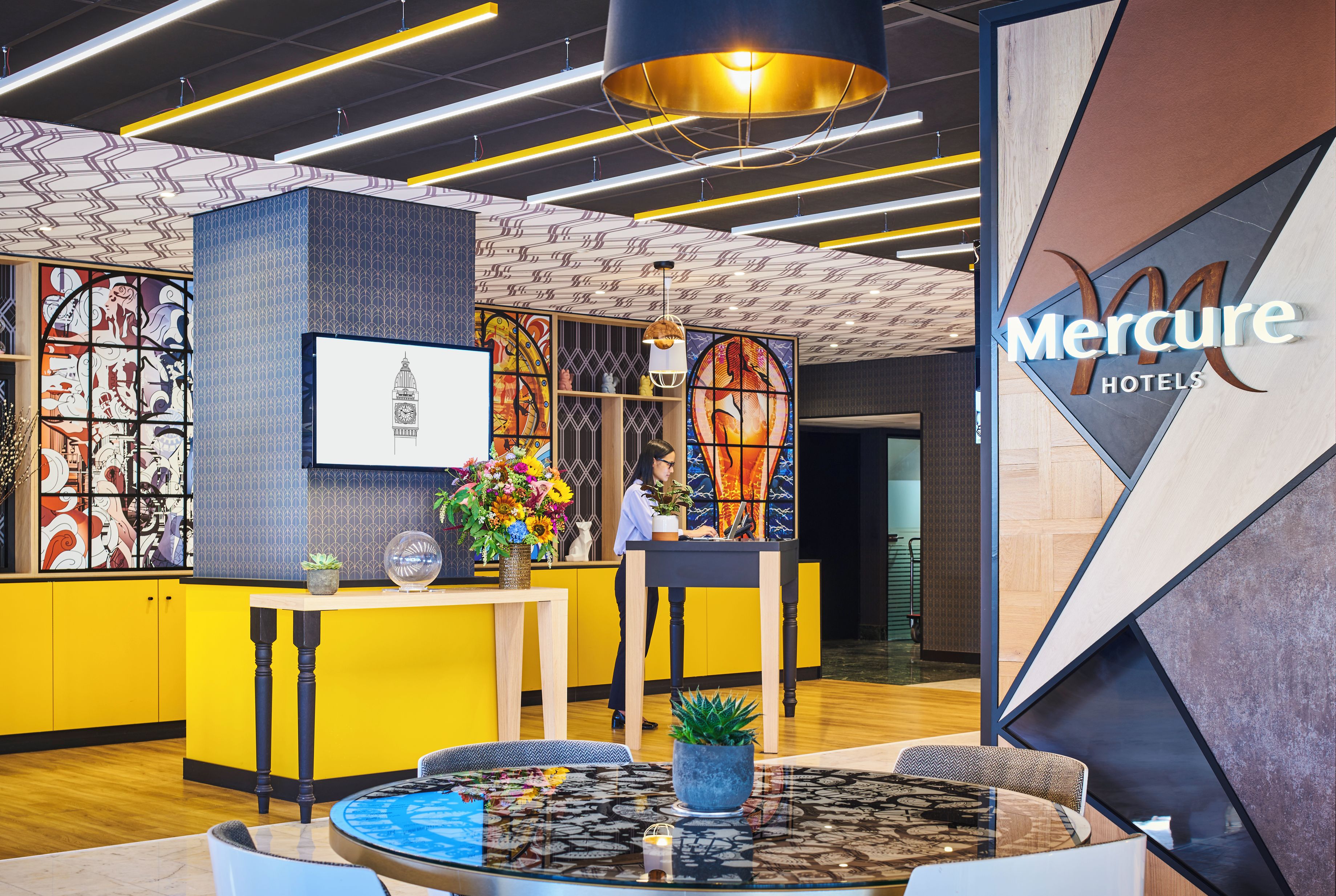 Zona de recepción de hotel con un diseño moderno, que incluye un mostrador de recepción, un reloj grande y una pantalla digital mostrando el nombre del hotel.