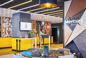 A hotel reception area with a modern design, including a reception desk, a large clock, and a digital screen displaying the hotel name.