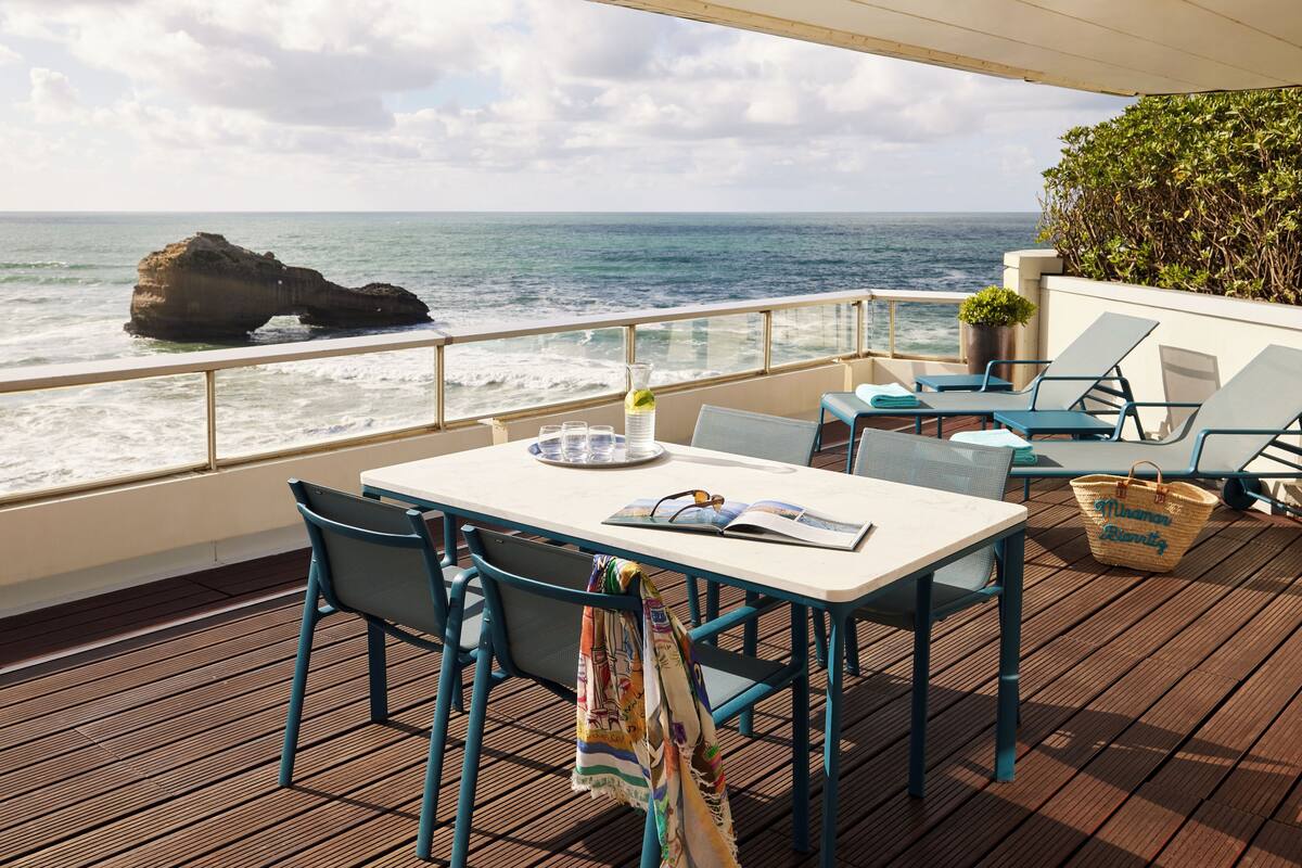 Una terraza con mesa y sillas, con vista al océano y una formación rocosa en la distancia.