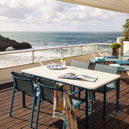 A deck with a table and chairs, overlooking the ocean with a rocky formation in the distance.