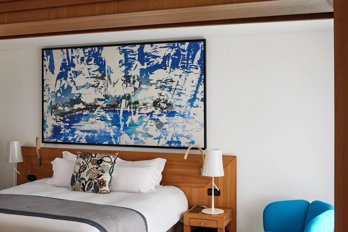 Una habitación de hotel con una cama grande, una mesita de noche, un sillón azul y un cuadro abstracto de gran tamaño en la pared.