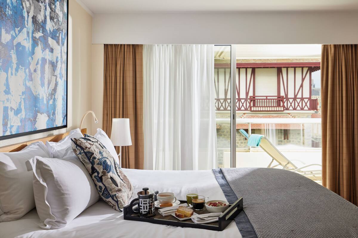 Una habitación de hotel con una cama, una bandeja con productos para el desayuno, vistas a un balcón con barandilla roja y un cuadro en la pared.