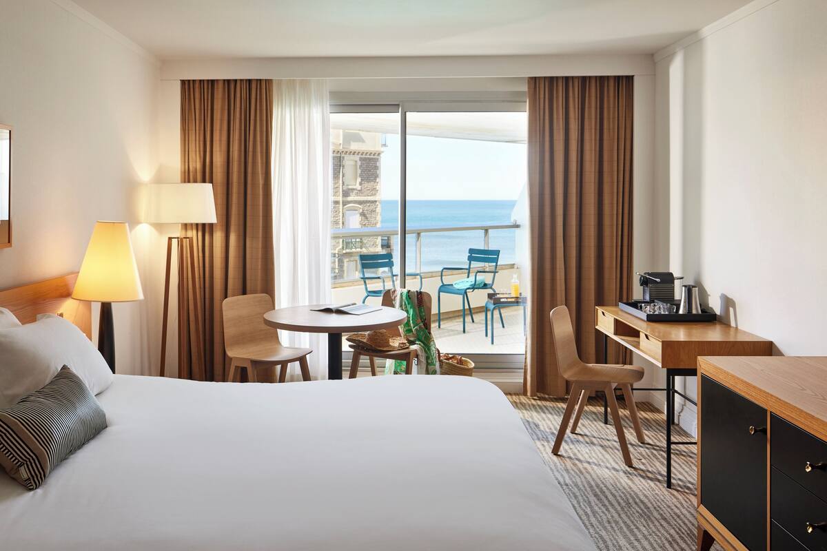 Habitación de hotel con una cama grande, un escritorio con cafetera, una silla, una mesita con un libro y un balcón con vistas al mar.