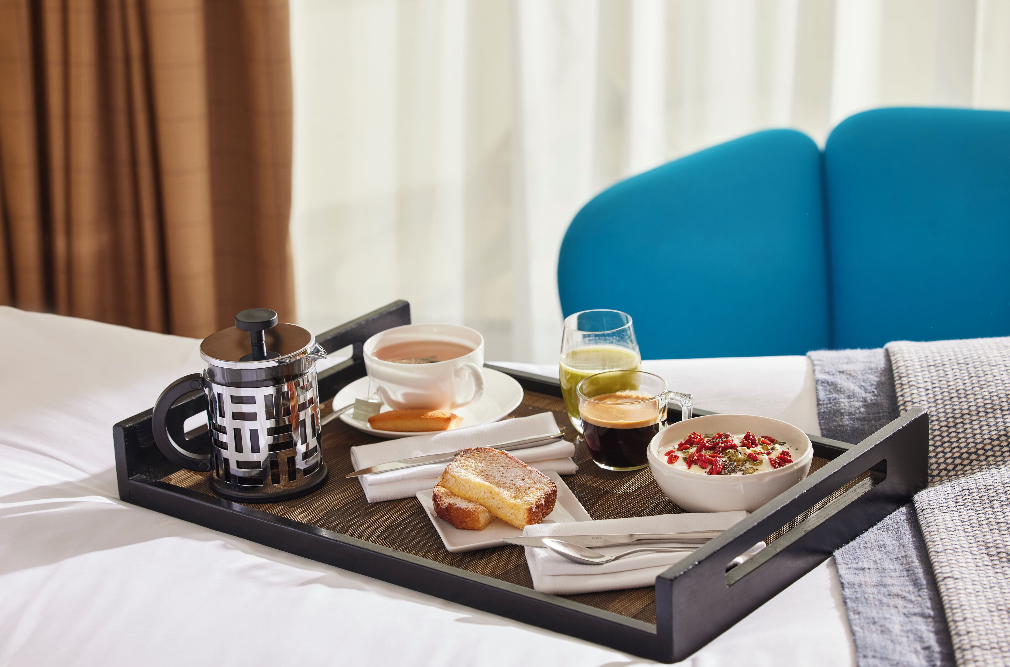 A breakfast tray on a bed with a cup of coffee, a glass of juice, a bowl of cereal, and a slice of toast.