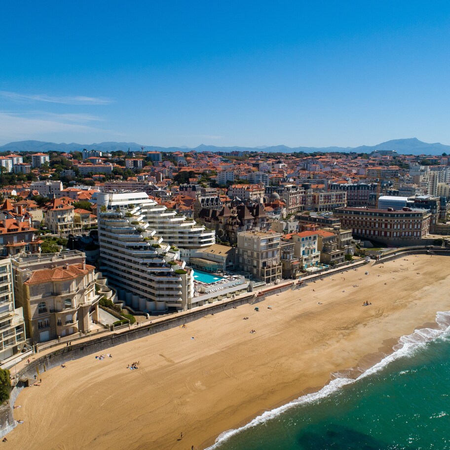 An aerial view of a coastal city with a beach, buildings, and mountains in the background.