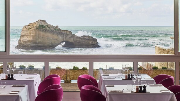 Restaurant amb taules preparades per dinar, grans finestres que ofereixen vista al mar i a una costa rocosa.