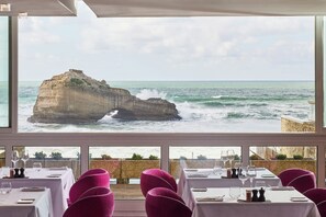Un restaurante con mesas dispuestas para comer, grandes ventanales que ofrecen vistas al mar y a la costa rocosa.